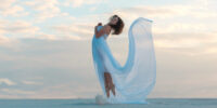 A girl in a fly white dress dances and poses in the sand desert at sunset.